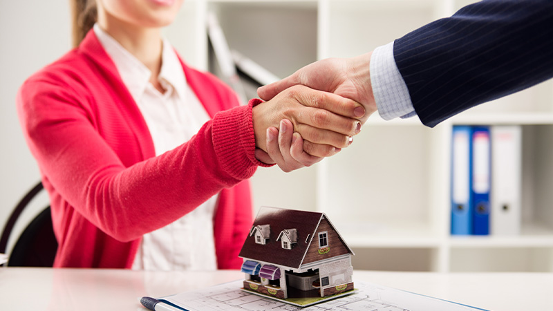 A real estate agent and a client shaking hands after buying deal with a model house on the table.